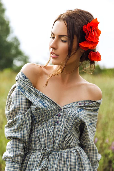 Outdoor portret van een mooie brunette vrouw in jurk met rode bloemen in haar haar in het veld. — Stockfoto