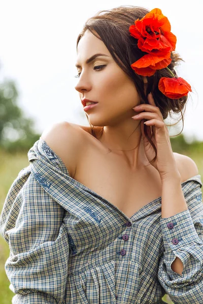 Outdoor portret van een mooie brunette vrouw in jurk met rode bloemen in haar haar in het veld. — Stockfoto