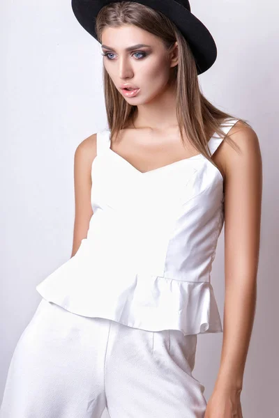 Girl in white clothes and black hat posing in studio on white background.