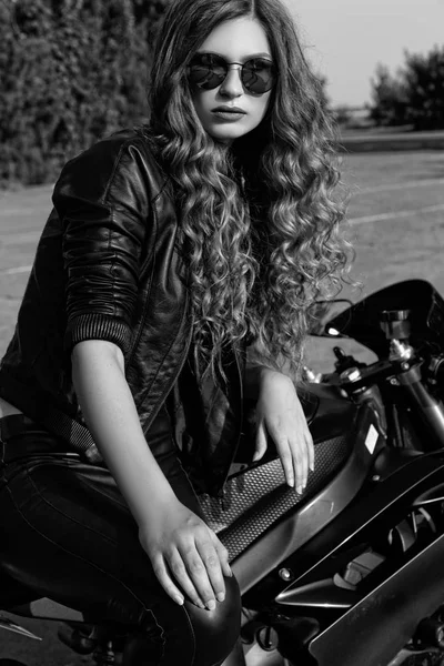 Jovem mulher em roupas de couro e óculos de sol perto de uma motocicleta. Foto em preto e branco — Fotografia de Stock
