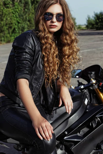 Jovem mulher em roupas de couro e óculos de sol perto de uma motocicleta — Fotografia de Stock