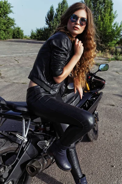Jovem mulher em roupas de couro e óculos de sol perto de uma motocicleta — Fotografia de Stock