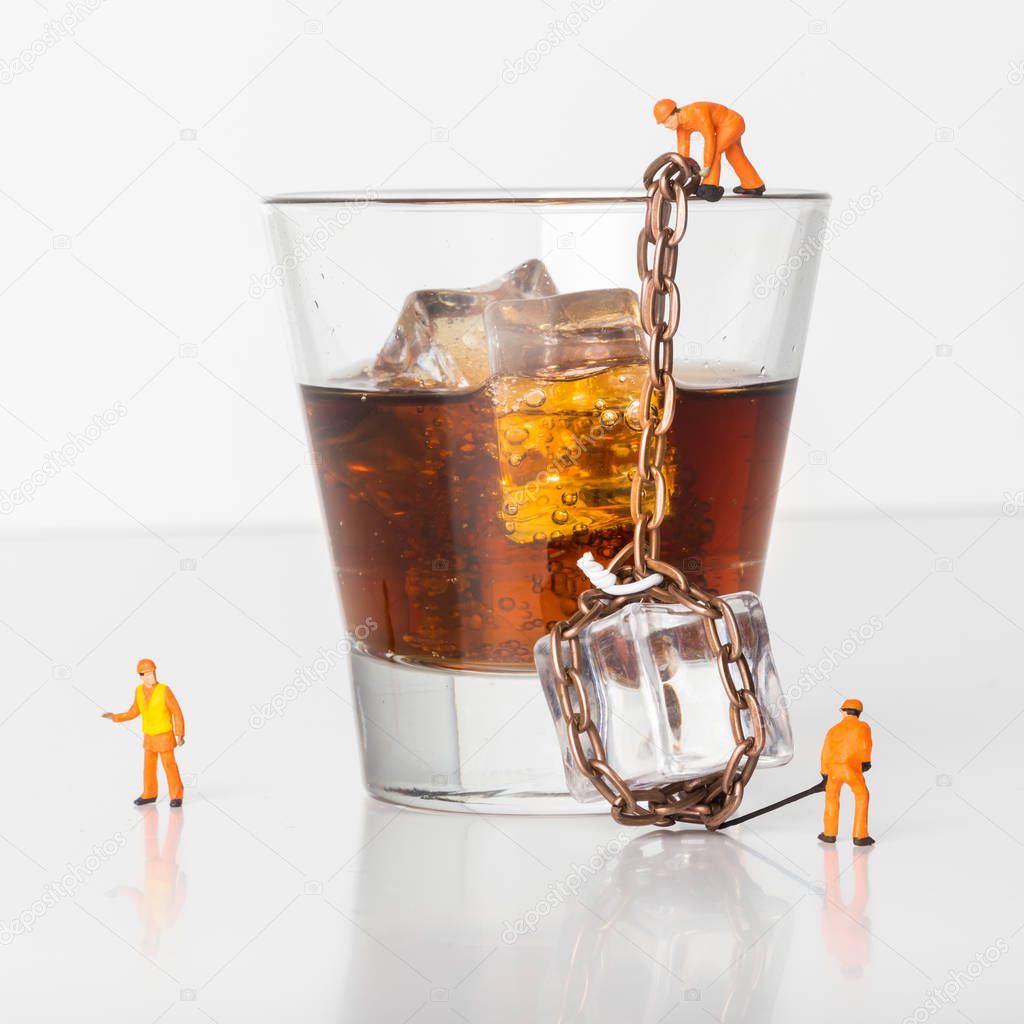Miniature people. The concept of alcohol in bars and restaurants. Miniature toy workers loaded an ice cube in a glass of brandy. Close-up view.