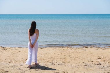Kumsalda ayakta ve denize bakarak yaz beyaz elbiseli genç esmer kadın.