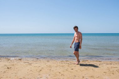 Sakallı mavi elbiseli bir adam sahilde yürüyor.