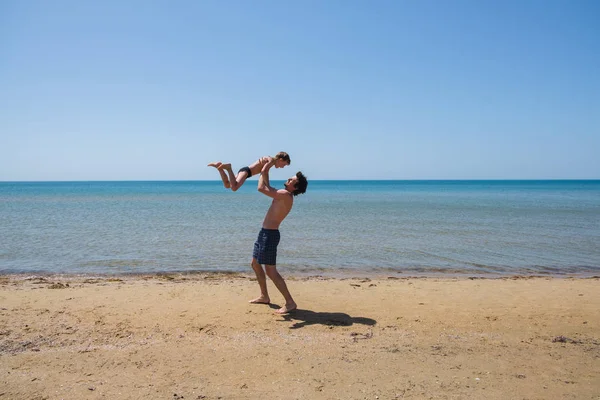 Młody szczęśliwy ojciec trzyma się w jego ramiona mały syn stawiania go na plaży. — Zdjęcie stockowe