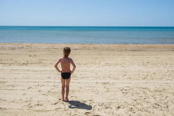Niño de seis años en bañador se para en la playa y mira al mar . — Foto de Stock