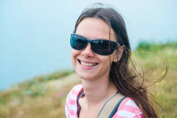Женщина 25-30 в солнечных очках стоит на холме. Портрет крупного плана. Брюнетка с длинными волосами, улыбающаяся . — стоковое фото