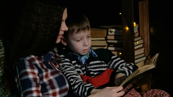 Moeder en zoon zitten in een stoel en het lezen van een boek gewikkeld in een deken. — Stockvideo