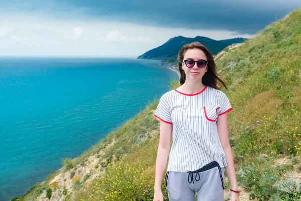 Mujer joven en gafas de sol se para en la colina y mira al frente del mar . — Foto de Stock