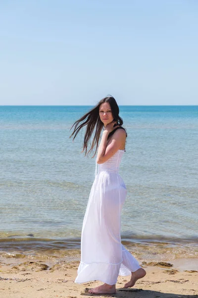 Mujer morena en un vestido blanco camina por la playa . — Foto de Stock