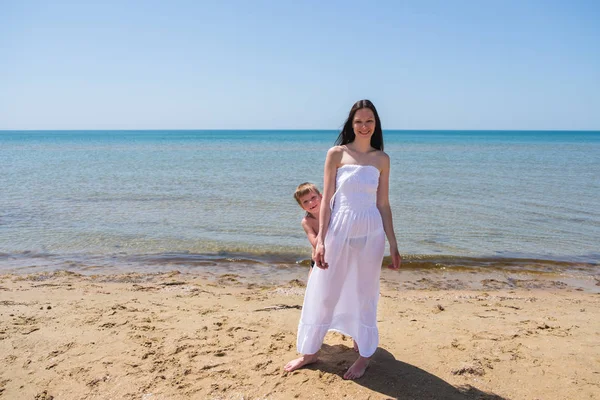 Morena en vestido largo y transparente con su hijo en la playa . — Foto de Stock