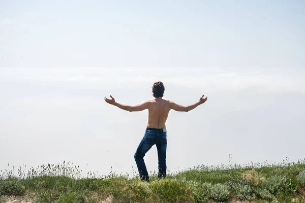 Junger Mann mit nacktem Oberkörper in Jeans und Brille steht am Rande einer Klippe und blickt in den Nebel. — Stockfoto