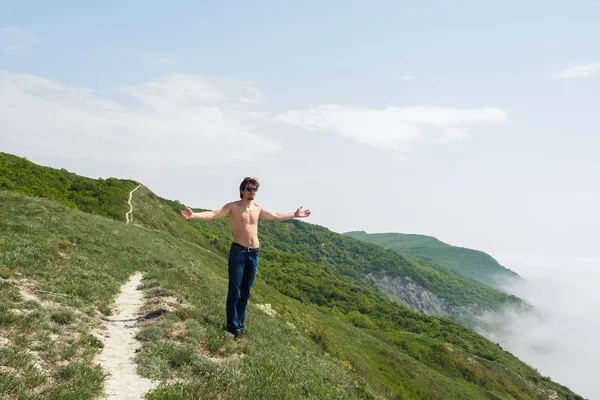 Joven con el torso desnudo en jeans y gafas está de pie en el borde de un acantilado y mira a la niebla . — Foto de Stock