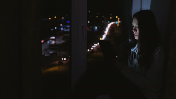 幸せな女は、タブレットを使用してビデオ チャットします。夜の暗闇の中で窓辺に座ってください。. — ストック動画