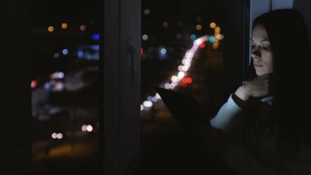 Frau, die in der dunklen Nacht auf der Fensterbank sitzt und auf die Straße blickt. Lesebuch im Tablet. — Stockvideo