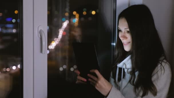 女性はタブレット コンピューターで映画を見て、笑っています。暗い夜に窓辺に座ってください。 — ストック動画