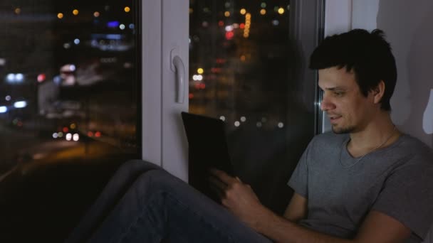 L'uomo è seduto sul davanzale della finestra nella notte buia e navigando web su tablet . — Video Stock