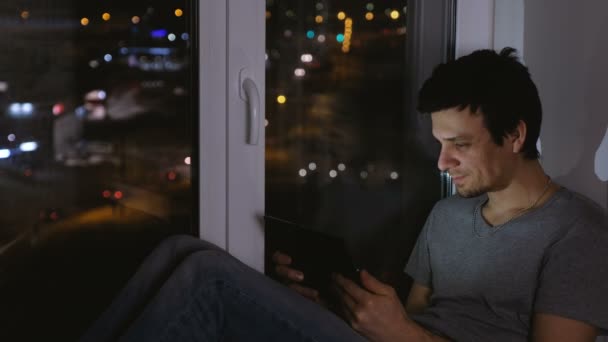 Człowiek ogląda film na telefon i śmiejąc się. Siedząc na parapecie w ciemną noc. — Wideo stockowe