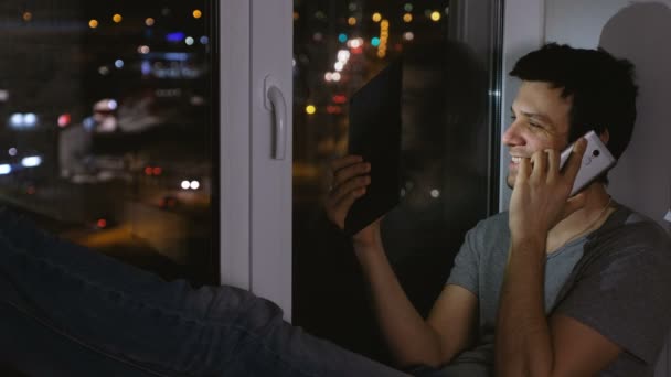 O homem feliz está a espetar o telemóvel e a ver tablet. Sentado no peitoril da janela à noite escura . — Vídeo de Stock