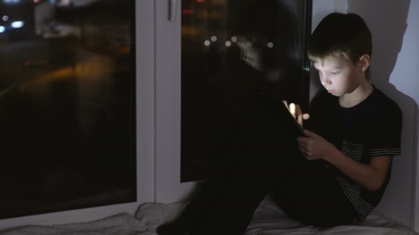 Niño de 7 años buscando algo en la tableta. Siéntate en el alféizar de la ventana en la oscuridad por la noche . — Vídeos de Stock
