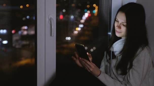 Femme naviguant sur le web sur un téléphone portable assis sur le rebord de la fenêtre dans la nuit noire. Fond routier . — Video