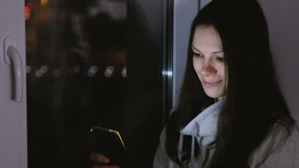 Mujer navegando por la web en el teléfono móvil sentado en el alféizar de la ventana en la noche oscura. Fondo de carretera — Vídeos de Stock