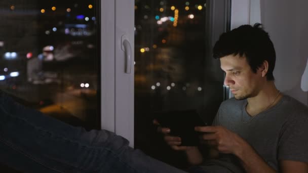 Hombre morena jugando juego en la computadora tableta. Sentado en el alféizar de la ventana en la oscuridad . — Vídeos de Stock