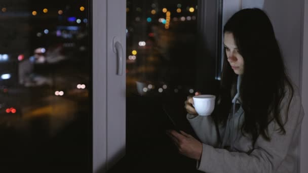 Mujer leyendo algo en la tableta y bebiendo té. Siéntate en el alféizar de la ventana en la oscuridad por la noche . — Vídeo de stock