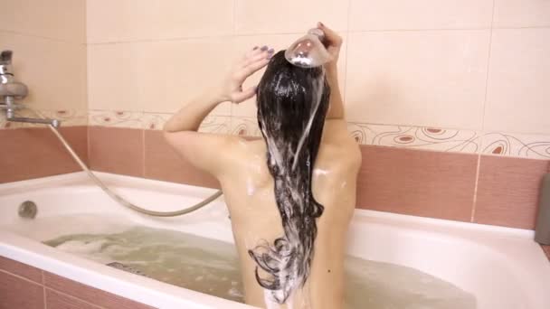 Неузнаваемая обнаженная брюнетка вымывает шампунь из волос, сидя в ванной с видом сзади — стоковое видео