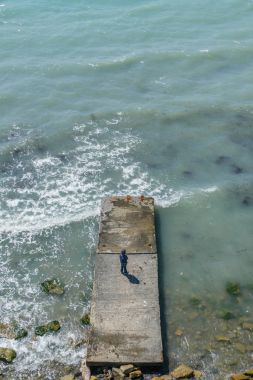 Altı yaşında çocuk ceket ve kot denizde iskelede bir fırtına sırasında dalgalar ile uzaktan görünümü duruyor