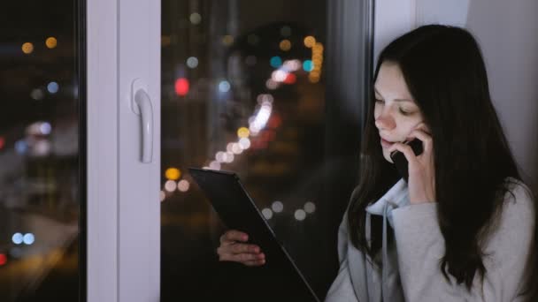 女人用手机聊天, 看着平板电脑。夜晚在黑暗中坐在窗台上. — 图库视频影像