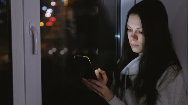 Kobieta, przeglądanie sieci web na telefon komórkowy siedząc na parapecie w ciemną noc. — Wideo stockowe