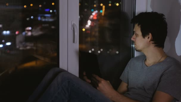 Hombre sentado en el alféizar de la ventana en la noche oscura y mirando a la calle. Uso de tableta para web . — Vídeos de Stock