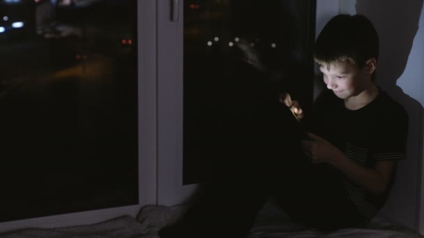 7ans garçon regardant quelque chose sur tablette et souriant. Asseyez-vous sur le rebord de la fenêtre dans le noir la nuit . — Video