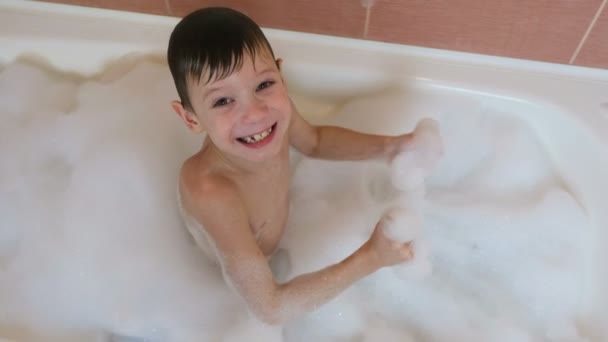 Niño de 6-7 años sentado en el baño con espuma mirando a la cámara y sonríe. Vista superior . — Vídeo de stock