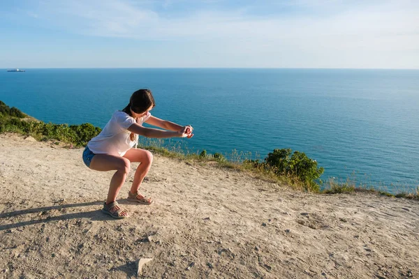 Woman brunette in light blouse and denim shorts doing squats on the mountain with seascape.