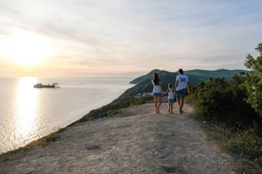 Aile anne, baba ve oğul günbatımında bir dağ deniz manzarası ile yürürken. Arkadan Görünüm.