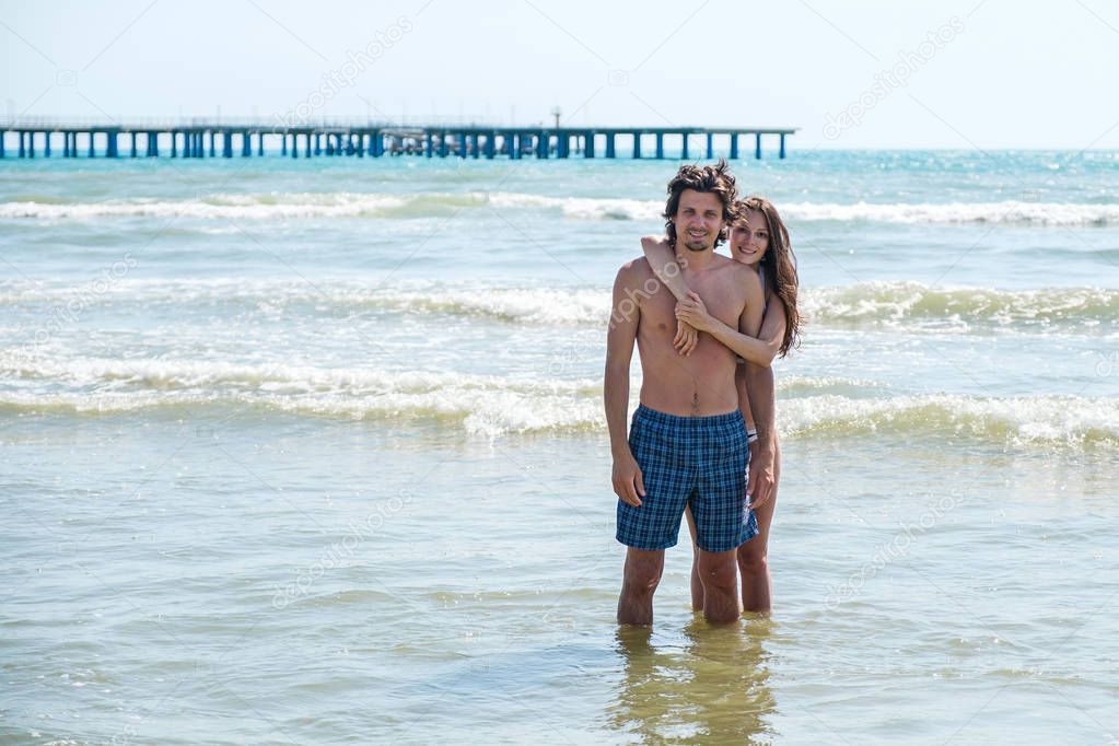Brunette woman in bikini hugs a beautiful long-haired man from behind, standing in the sea.
