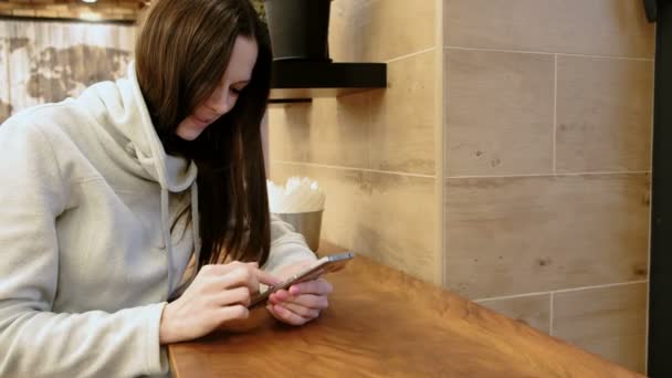 Souriant jeune femme brune dans une chemise lumineuse regardant dans son téléphone portable assis dans un café . — Video