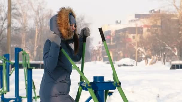 Frau in blauer Daunenjacke mit Kapuze im Skisimulator in einem Winterpark in der Stadt. — Stockvideo