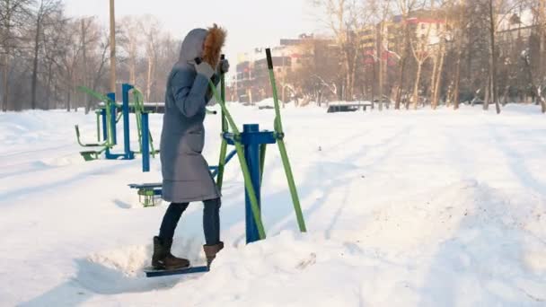 Frau in blauer Daunenjacke mit Kapuze im Skisimulator in einem Winterpark in der Stadt. — Stockvideo