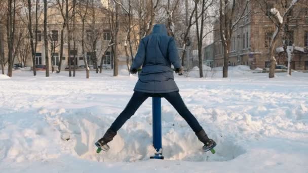 Mulher de casaco azul para baixo com um capuz está fazendo exercícios de pernas no simulador em um parque de inverno na cidade . — Vídeo de Stock