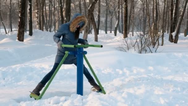 女人在蓝色羽绒服与胡德是做腿演习在模拟器在冬季公园前视图 — 图库视频影像