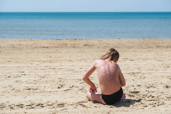 Blond dziecko na sobie czarne spodnie, siedząc na plaży. — Zdjęcie stockowe