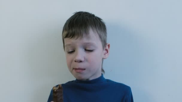 Niño de siete años con una chaqueta azul muerde y mastica una barra de chocolate. Mira la barra de chocolate — Vídeos de Stock