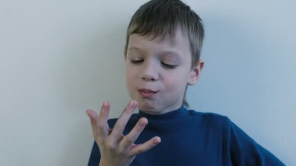 Primer plano niño de siete años en una camisa azul con una cara de chocolate lame sus dedos . — Vídeo de stock