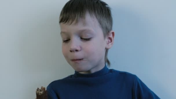 Niño de siete años con camisa azul se pone un chocolate entero en la boca y lo mastica . — Vídeos de Stock