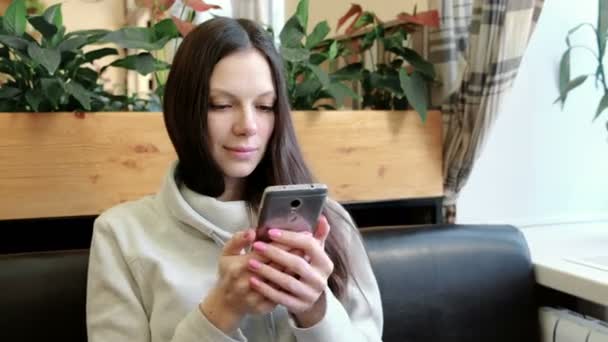 Jovem morena feliz está considerando algo interessante e jogando em seu celular no café — Vídeo de Stock