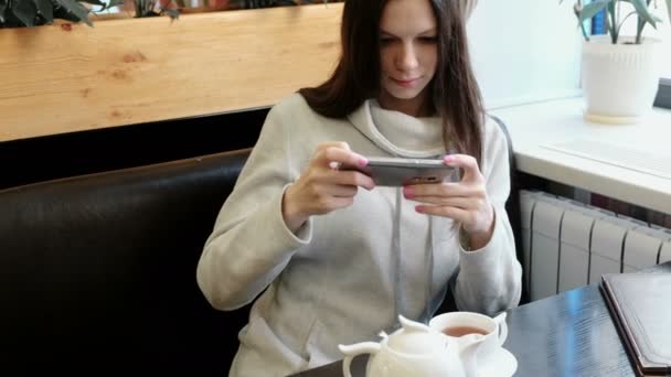 Genç kadın esmer resimleri çaydanlık ve bardak cep telefonunda bir kafede. Önden Görünüm. — Stok video